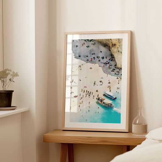 Framed artwork in a natural wood frame, leaning against a light-colored wall on a wooden table, depicting a watercolor-style beach scene with two boats and people on the shore.