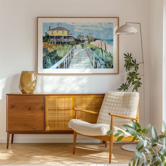 Framed beach house painting displayed above a mid-century modern cabinet in a sunlit room. The artwork depicts a boardwalk leading past a yellow stilt house and other colorful cottages toward a sandy shoreline, with tall grasses and a calm blue ocean in the distance.