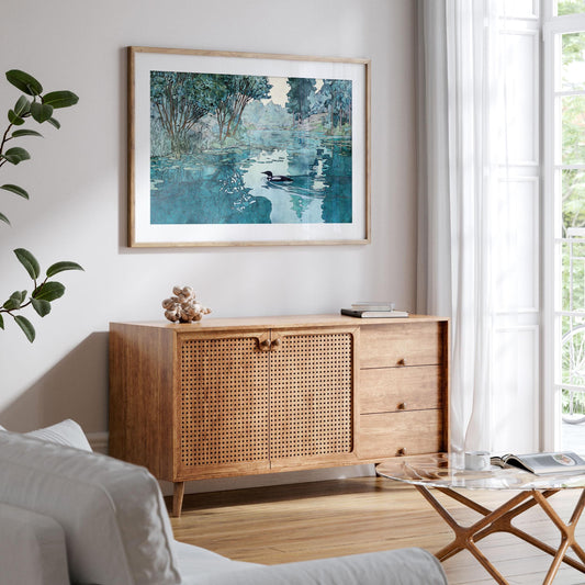 Framed art print of a loon swimming on a reflective lake, surrounded by trees and lily pads, displayed above a mid-century wooden sideboard in a bright, modern living room with natural light, a glass coffee table, and white curtains.