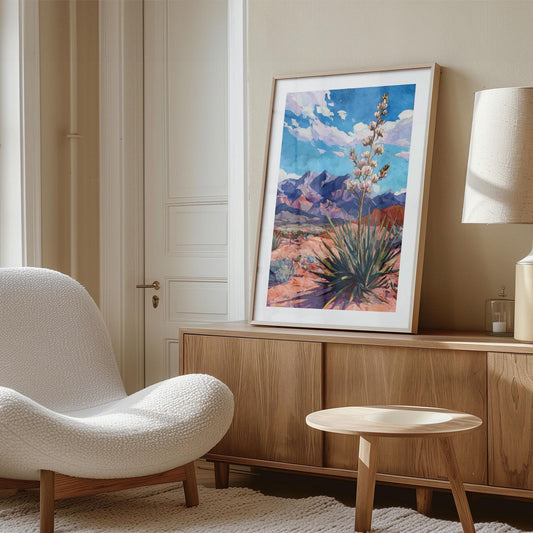Framed desert landscape painting featuring a blooming yucca plant in front of purple mountains under a vivid blue sky, displayed on a wooden sideboard in a modern living room. The space includes a textured white lounge chair, a round wooden side table, and a beige lamp, with soft natural light creating a warm, minimalist atmosphere.