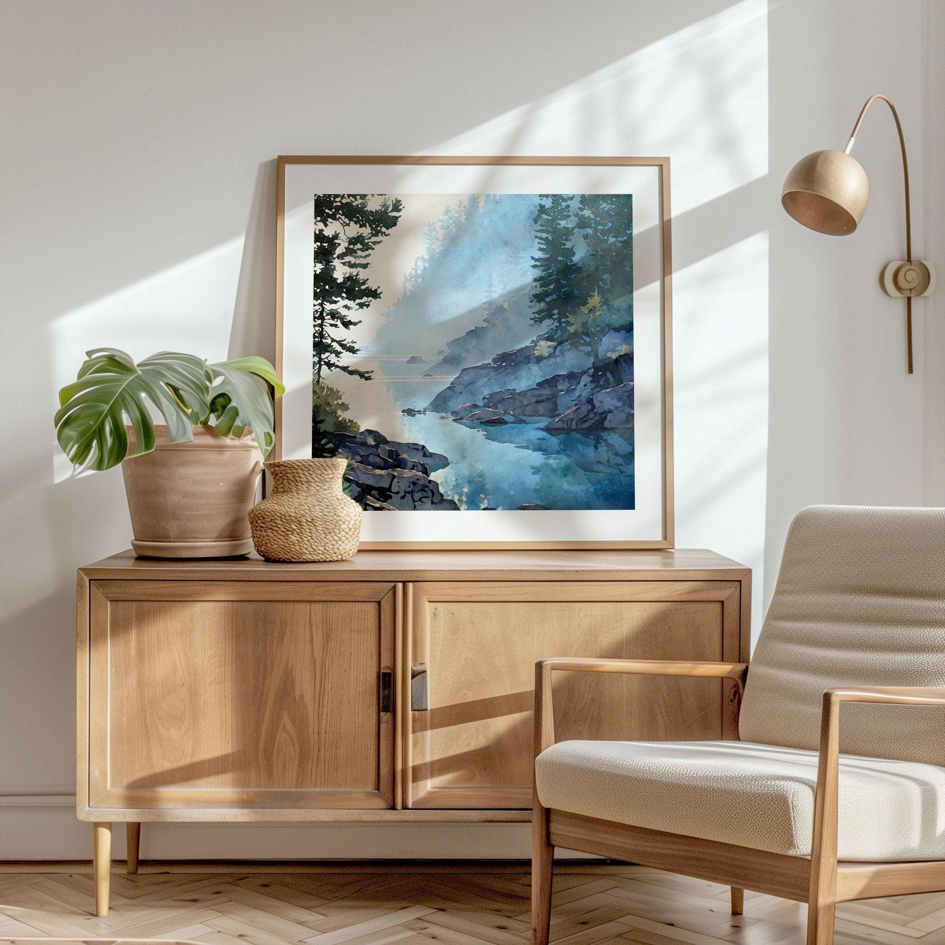 Styled interior photo featuring a framed watercolor-style landscape art print resting on a wooden cabinet. The artwork shows a misty blue lake with rocky shoreline and evergreen trees reflected in still water. Warm sunlight casts soft shadows across a neutral wall, with a modern chair, potted plant, and minimalist lamp creating a calm, Scandinavian-inspired home decor scene.