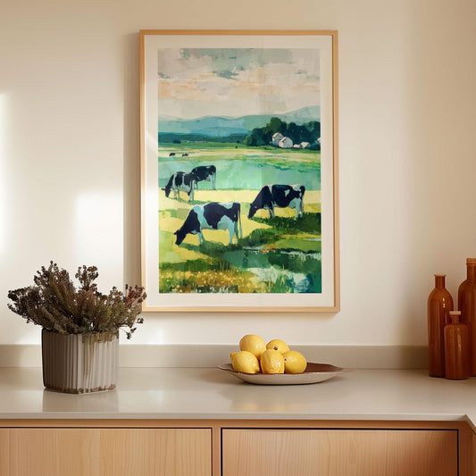Framed farmhouse-style painting of black and white cows grazing in a green meadow with distant hills and a cluster of barns. The artwork, done in bold impressionist brushstrokes, hangs on a neutral kitchen wall above a countertop decorated with a bowl of lemons, ceramic bottles, and a potted plant, creating a warm rustic-modern look.