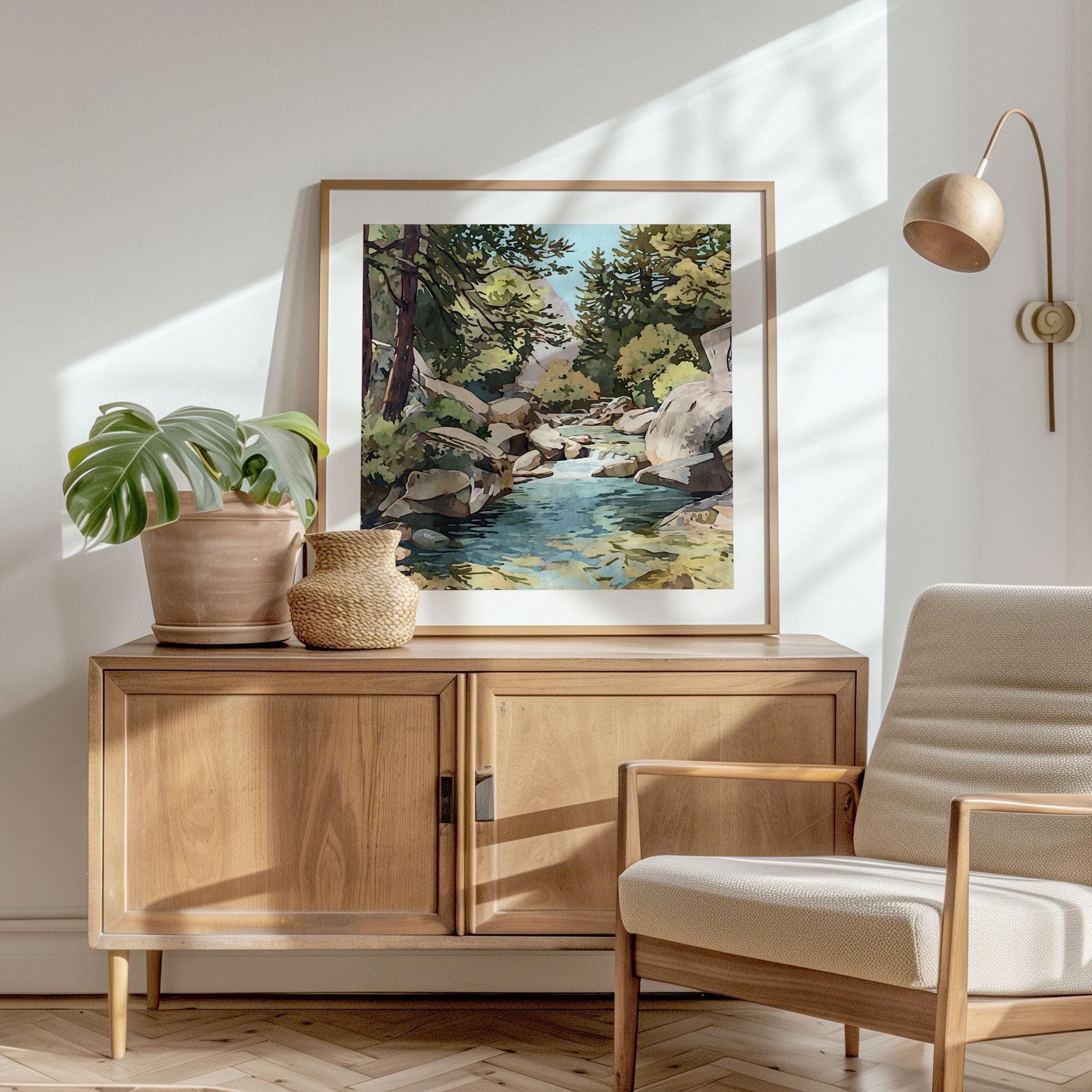 Framed painting of a rocky stream with pine trees and mountains displayed on a wooden sideboard, styled with a potted plant, woven vase, and modern chair in a sunlit, minimalist living space.