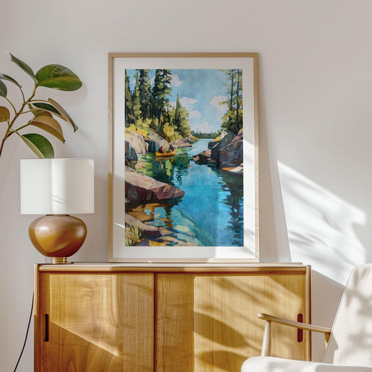 Framed painting of a canoe on a calm blue lake between pine-covered cliffs, displayed above a wooden cabinet with a modern lamp and houseplant in a sunlit room with minimalist decor.