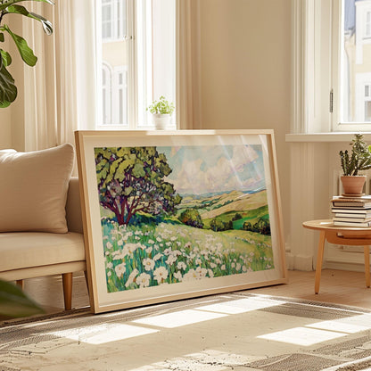 Framed painting of a bright meadow filled with white wildflowers and a large tree on the left, overlooking rolling green hills under a blue sky with fluffy clouds, leaning against a wall in a sunlit living room beside a beige sofa and small wooden table with plants and books.