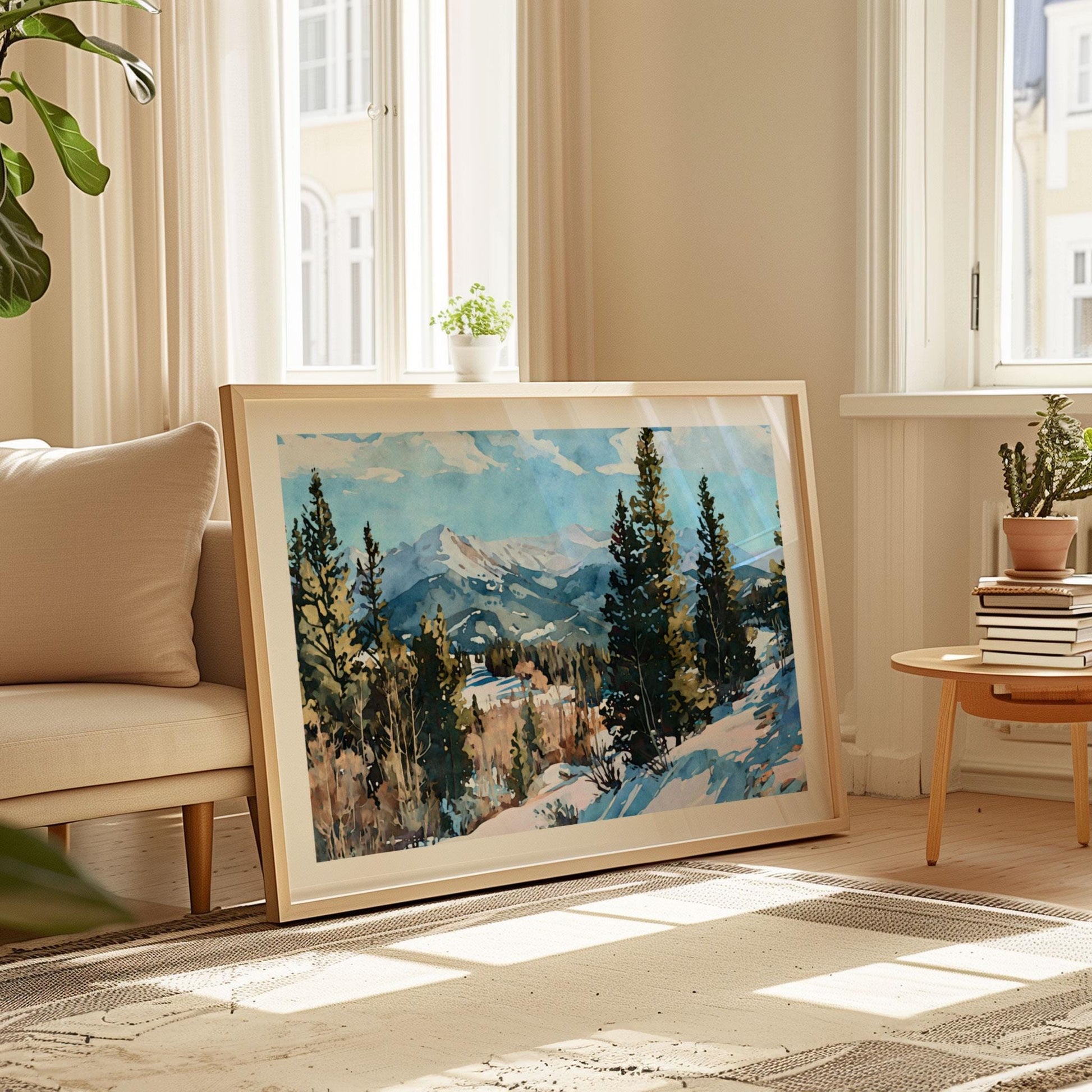 Framed watercolor painting of a snowy mountain landscape with evergreen trees, displayed leaning against a wall in a sunlit living room with neutral decor, beige sofa, potted plants, and a stack of books on a wooden side table.