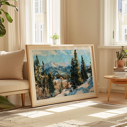 Framed watercolor painting of a snowy mountain landscape with evergreen trees, displayed leaning against a wall in a sunlit living room with neutral decor, beige sofa, potted plants, and a stack of books on a wooden side table.