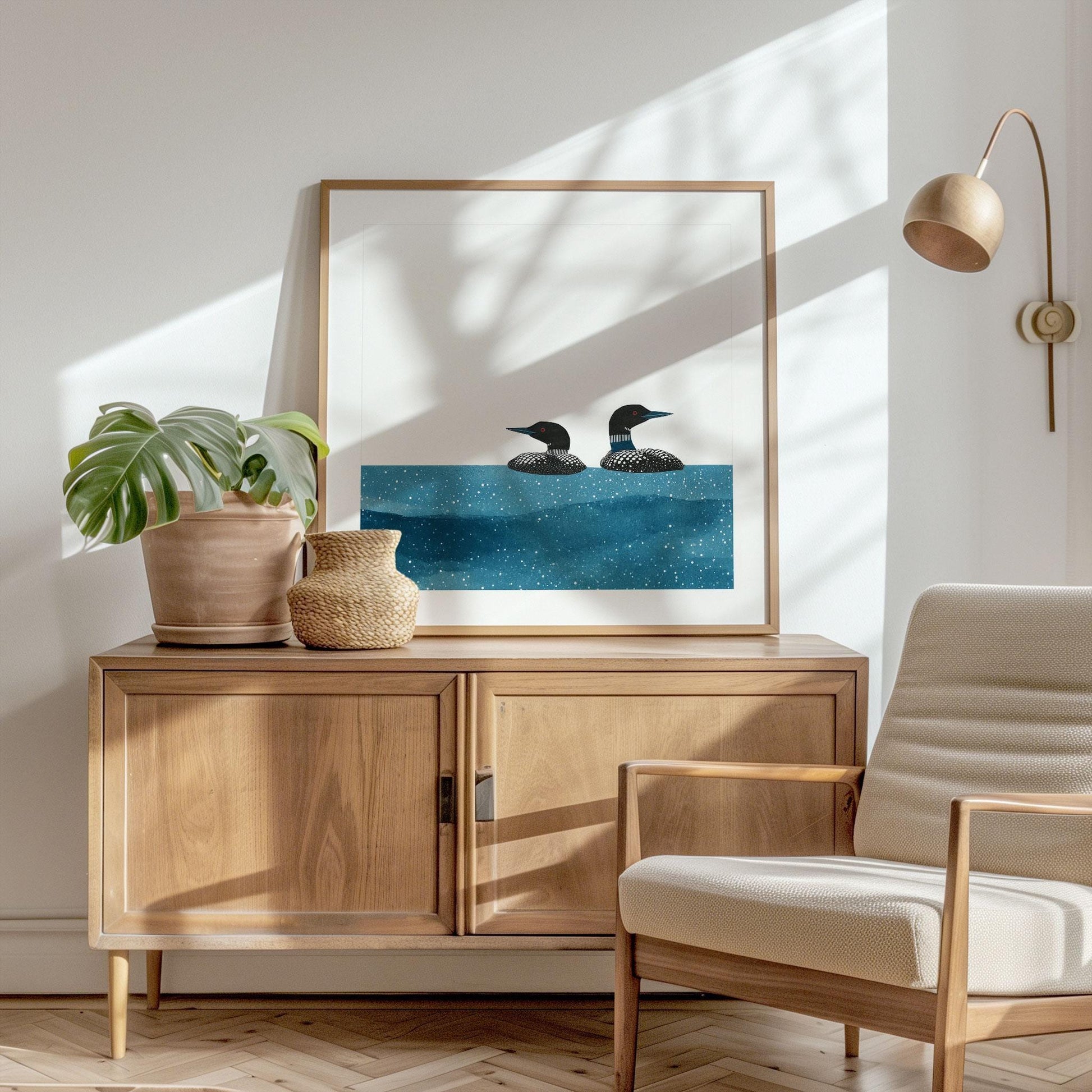 Minimalist loon art print in a light wood frame rests on a modern wooden sideboard. The artwork shows two black-and-white loons with red eyes gliding across textured blue water against a white background. Styled with a potted monstera plant, woven vase, neutral armchair, and brass wall lamp, the scene is filled with soft natural light and leafy shadows, creating a cozy, Scandinavian-inspired interior.