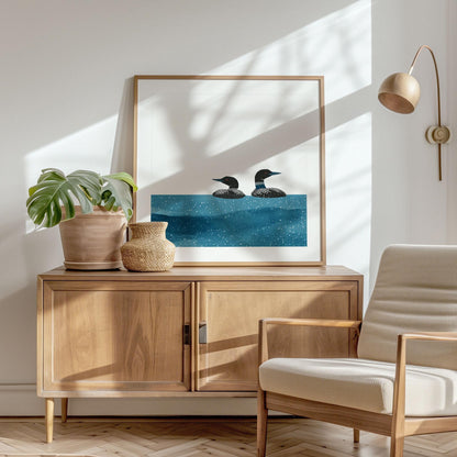Minimalist loon art print in a light wood frame rests on a modern wooden sideboard. The artwork shows two black-and-white loons with red eyes gliding across textured blue water against a white background. Styled with a potted monstera plant, woven vase, neutral armchair, and brass wall lamp, the scene is filled with soft natural light and leafy shadows, creating a cozy, Scandinavian-inspired interior.