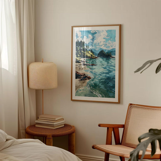 Framed coastal landscape art print hanging on a beige wall in a cozy bedroom with minimalist decor. The artwork depicts turquoise waves, a sandy beach, rocky cliffs, and evergreen trees under a bright sky with clouds. The room features a woven chair, a round wicker side table with stacked books, and a textured beige lamp beside soft curtains.