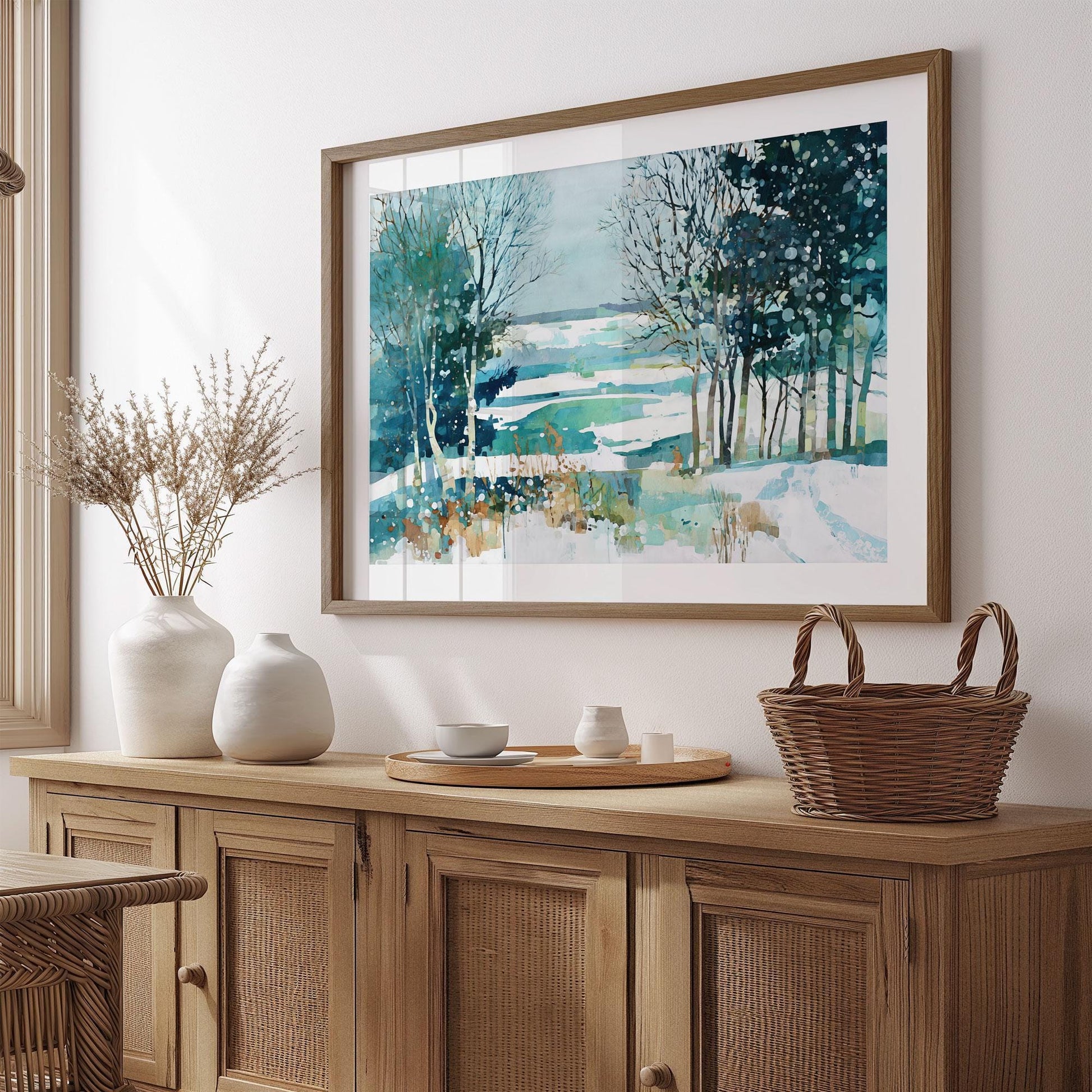 Framed watercolor painting of a snowy winter landscape with trees and a frozen lake in teal, blue, and white tones, displayed above a wooden sideboard with ceramic vases, a tray, and a woven basket in a cozy, minimalist room.