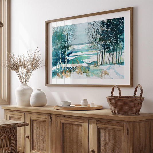Framed watercolor painting of a snowy winter landscape with trees and a frozen lake in teal, blue, and white tones, displayed above a wooden sideboard with ceramic vases, a tray, and a woven basket in a cozy, minimalist room.
