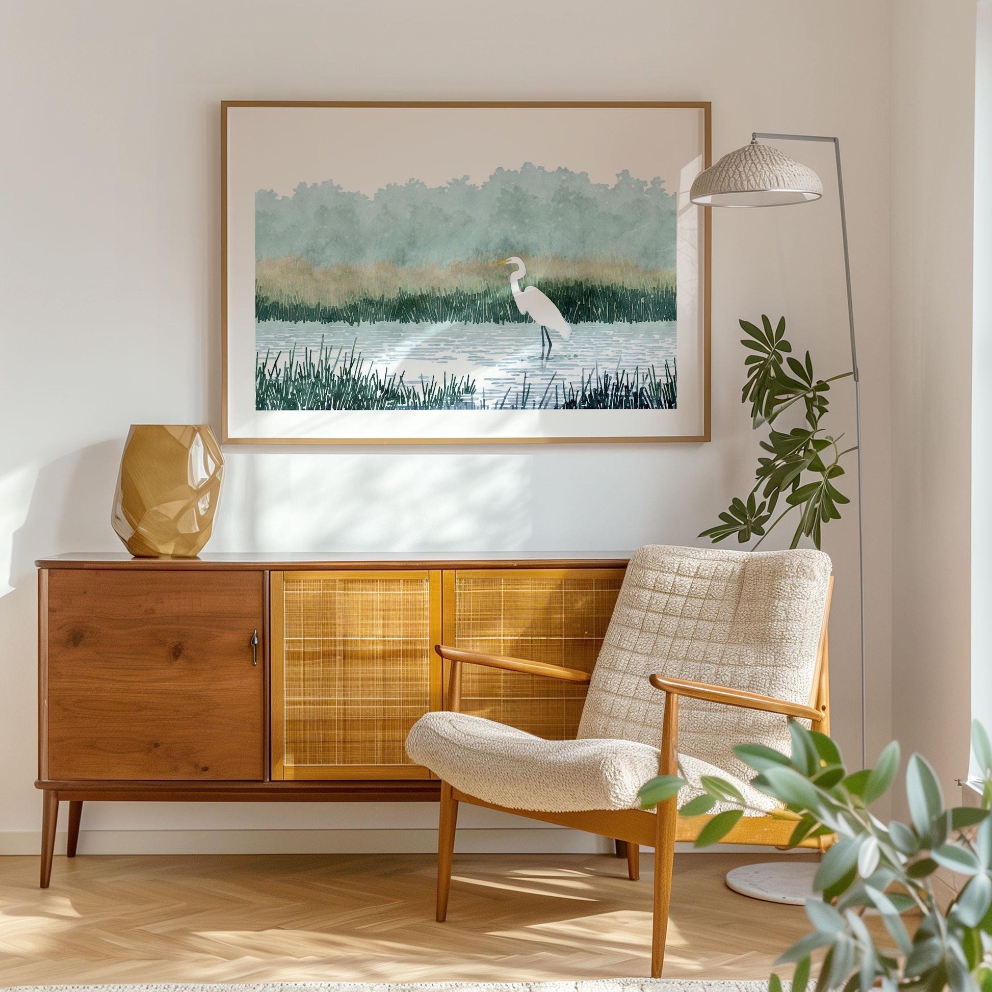 Framed watercolor-style print of a white heron standing in a marsh, hung above a mid-century modern sideboard in a cozy living room with a textured cream armchair, floor lamp, and leafy green plant.