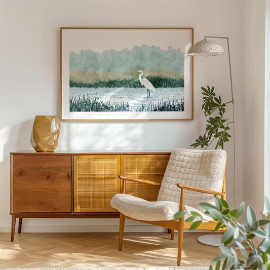 Framed watercolor-style print of a white heron standing in a marsh, hung above a mid-century modern sideboard in a cozy living room with a textured cream armchair, floor lamp, and leafy green plant.