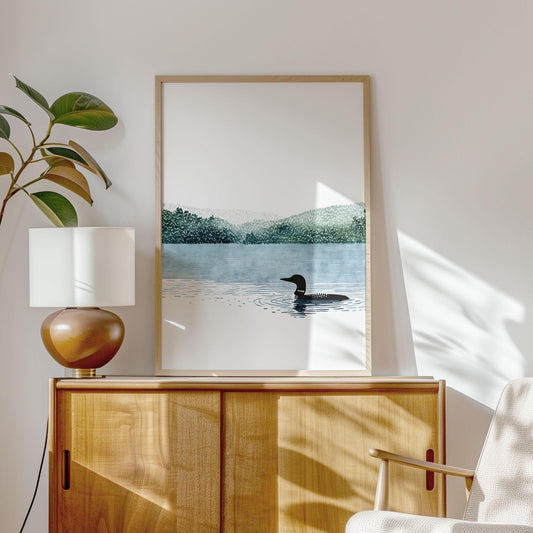 Framed minimalist watercolor art print displayed on a light wood sideboard in a bright, modern interior. The artwork shows a solitary loon swimming on a calm blue lake with soft green forested hills in the distance and a wide white sky. Sunlight casts gentle shadows across the wall, with a potted plant and table lamp beside the frame, creating a serene, nature-inspired home setting.