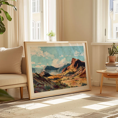 Large framed painting of a desert canyon with red rock cliffs, rolling hills, and distant blue mountains under a bright sky with scattered clouds, resting on the floor in a sunlit living room beside a cream sofa, potted plants, and a small table stacked with books.