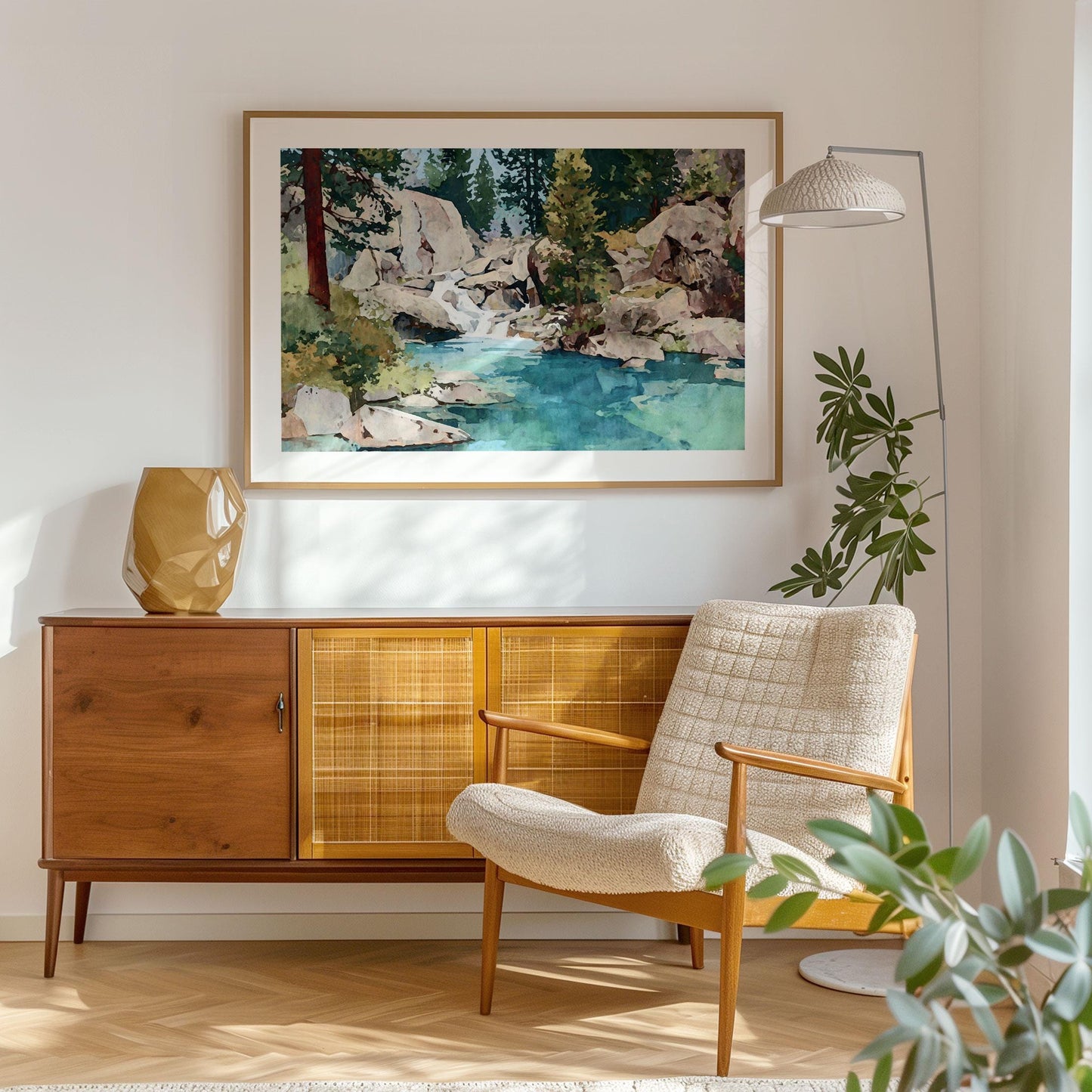 Framed watercolor landscape art print of a rocky mountain stream with turquoise water and pine trees, hanging above a mid-century modern wood credenza. The scene is softly lit by natural light, accompanied by a beige armchair, geometric gold vase, and a tall floor lamp, creating a warm, minimalist living room setting with cozy and organic textures.