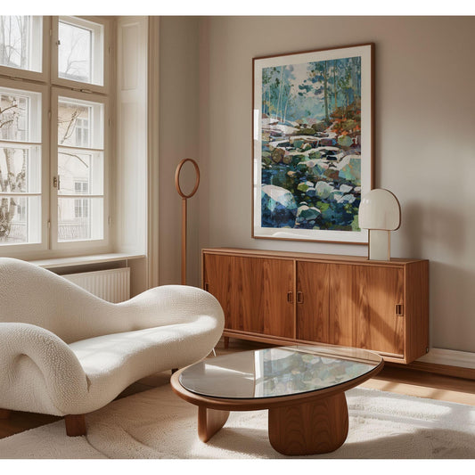 Modern living room with mid-century and Scandinavian influences, featuring a framed impressionist forest stream painting in cool blues and greens above a walnut credenza. The space includes a sculptural white boucle sofa, a glass-top wood coffee table, and a minimalist lamp, all bathed in soft natural light from large windows.