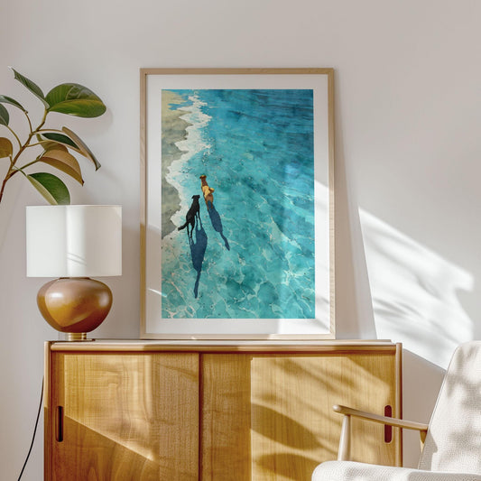 Framed art print of two dogs walking side by side along the ocean’s edge, their shadows stretching across shimmering turquoise water. Displayed in a bright, modern interior with warm wood accents, the watercolor artwork evokes calm, sunshine, and the joy of a beachside stroll.