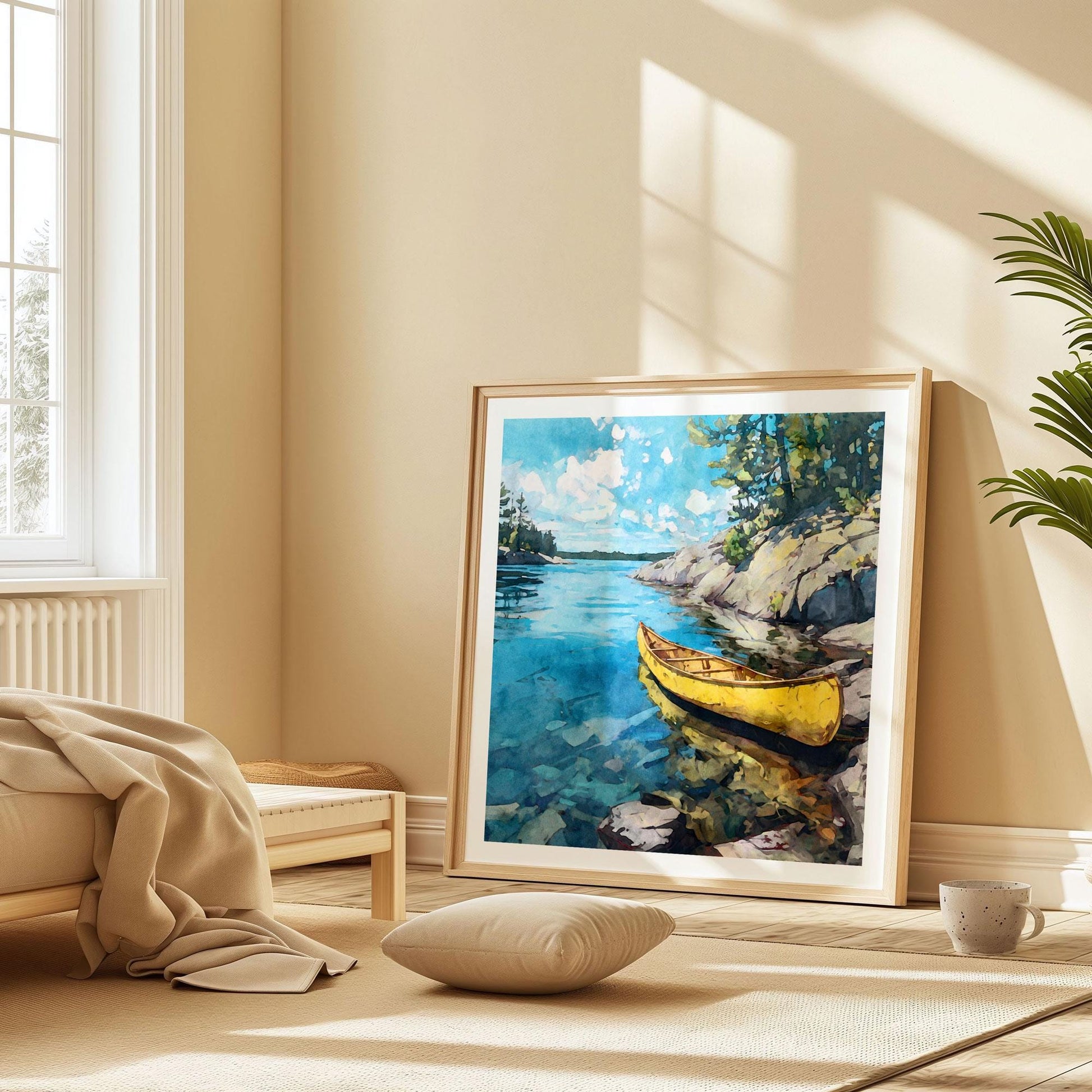 Large framed watercolor-style print of a yellow canoe on a clear blue lake leans against a wall in a warm, sunlit bedroom. The serene artwork shows rocky shoreline, pine trees, and bright sky reflected in calm water. A neutral bed, soft blanket, floor cushion, and a mug create a cozy, minimalist setting with natural light streaming through a nearby window.
