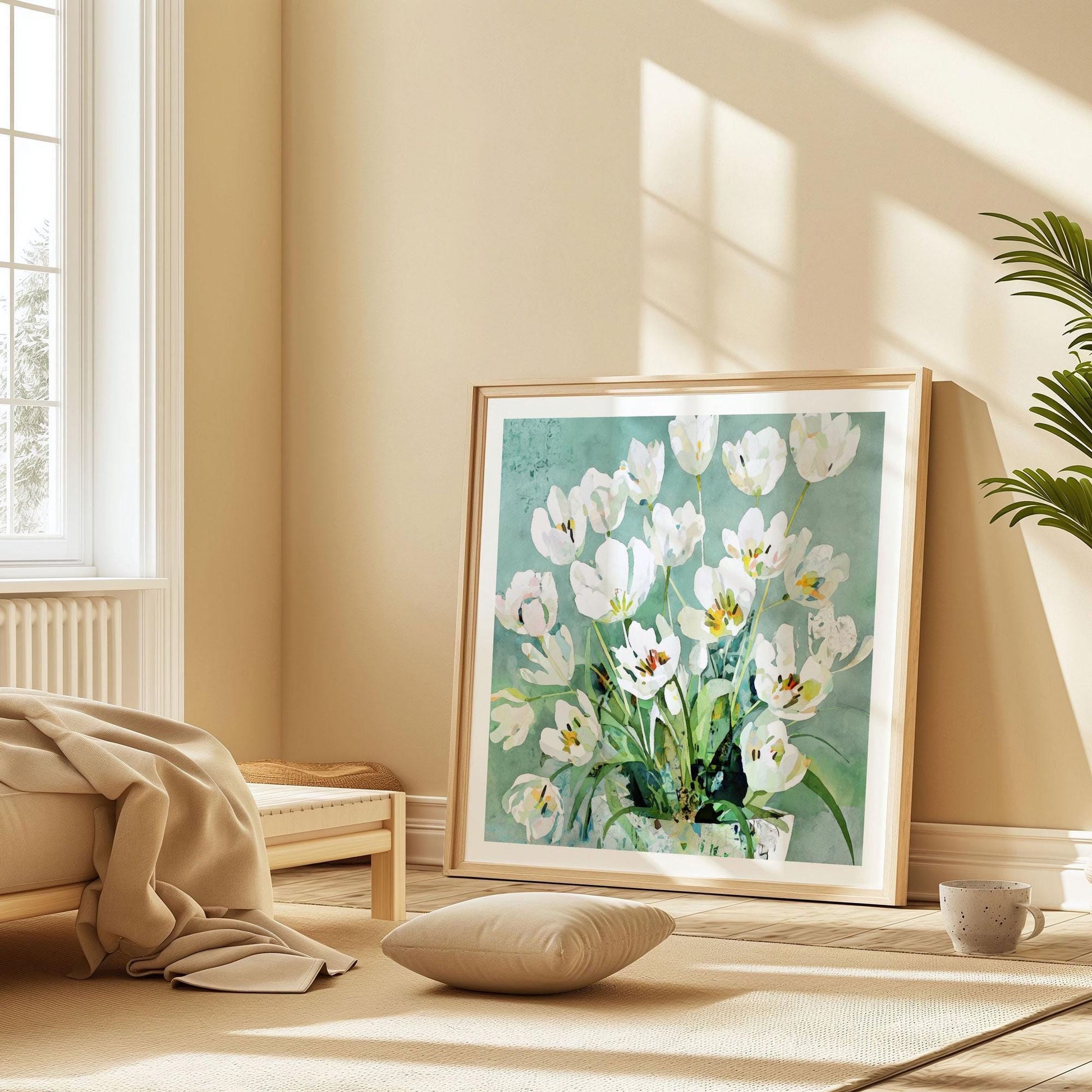 Framed art print of white tulips in a blue-green vase rests on the floor of a cozy sunlit room with beige walls and minimal decor. The watercolor-style floral artwork features soft teal and green tones. A cushion, beige throw blanket, and potted plant create a warm, serene atmosphere beside a large window.