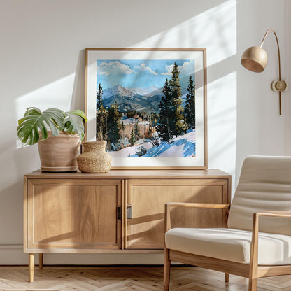 Framed watercolor painting of a snowy mountain landscape with evergreen trees, displayed on a wooden cabinet in a bright, modern room with neutral tones, a potted monstera plant, woven vase, and a cozy armchair beside a brass wall lamp.