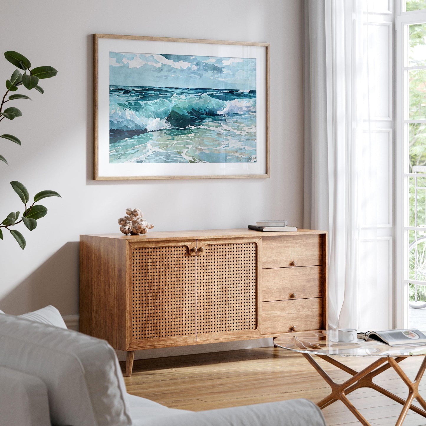 Framed watercolor painting of turquoise ocean waves hangs above a mid-century modern wooden cabinet in a bright living room with light wood floors, white walls, a glass coffee table, and large windows with sheer curtains.