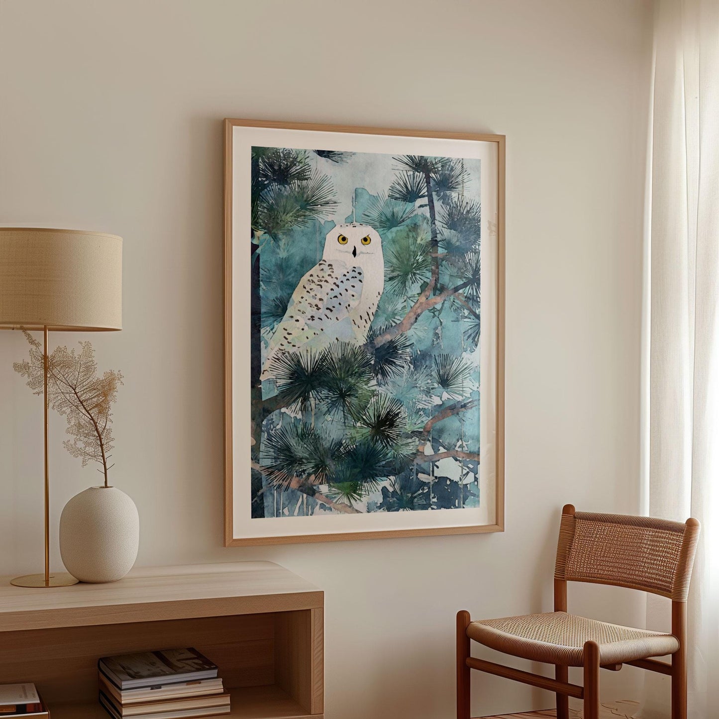 Framed watercolor-style art print of a snowy owl perched in pine branches, featuring cool blue and green tones with the owl’s bright yellow eyes. Displayed in a minimalist room with neutral walls, a woven chair, a wooden console with books, a beige lamp, and soft natural light filtering through sheer curtains.