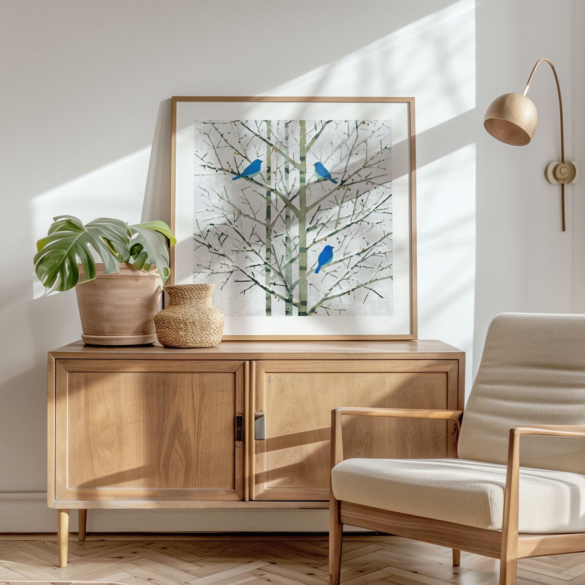 Framed print of three bluebirds perched on bare winter branches, styled in a bright Scandinavian-inspired room. The artwork rests on a mid-century wood sideboard beside a terracotta planter with a monstera leaf and a woven vase. Sunlight streams across a cream armchair, herringbone wood floor, and white wall, creating a warm, minimalist atmosphere.