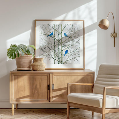 Framed print of three bluebirds perched on bare winter branches, styled in a bright Scandinavian-inspired room. The artwork rests on a mid-century wood sideboard beside a terracotta planter with a monstera leaf and a woven vase. Sunlight streams across a cream armchair, herringbone wood floor, and white wall, creating a warm, minimalist atmosphere.