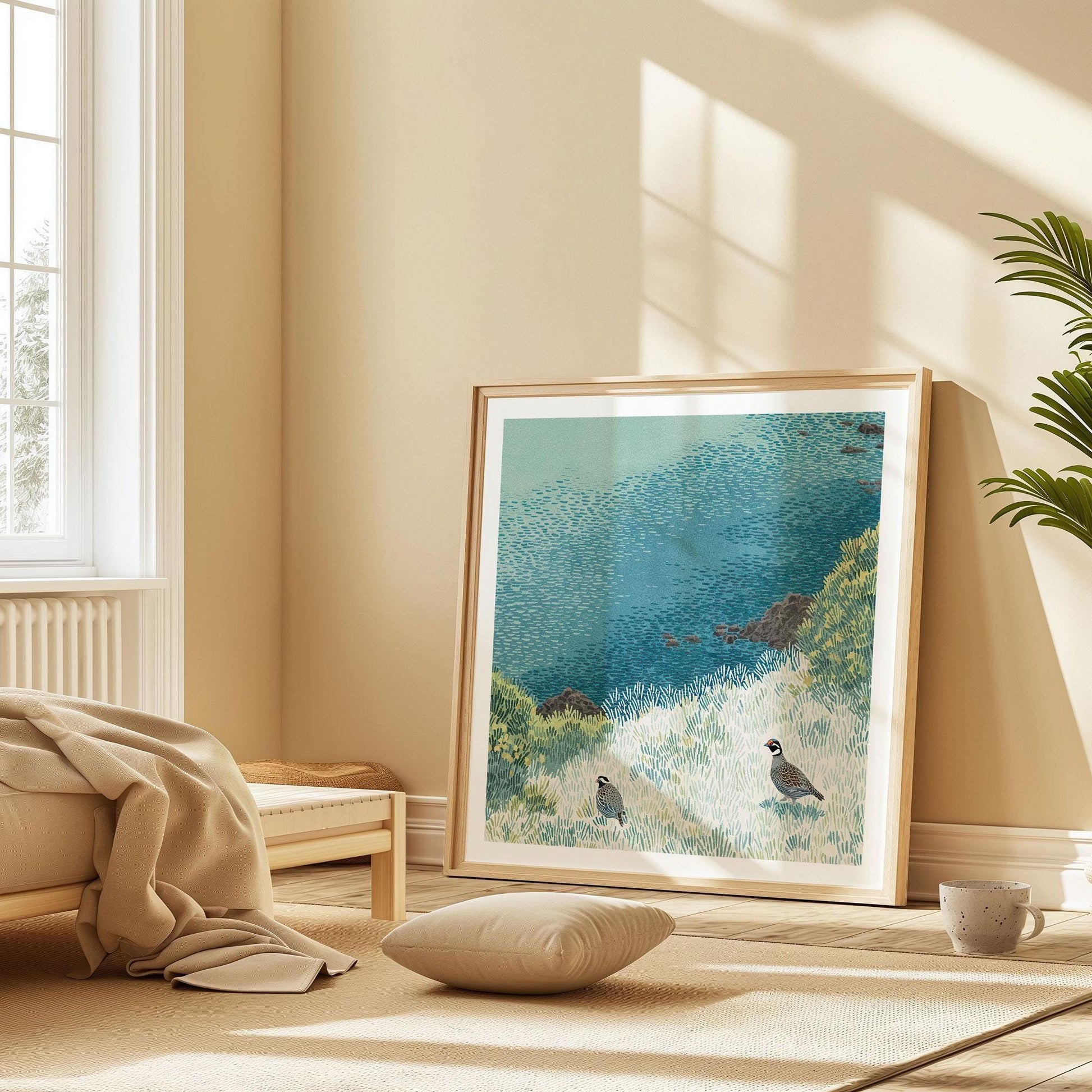 Framed art print of two California quail in a coastal meadow, propped against a warm beige wall in a minimalist sunlit room. The illustration features textured grasses, rocky outcrops, and a rich blue sea, adding a tranquil, nature-inspired accent to the cozy neutral space with its low furniture and soft natural light.