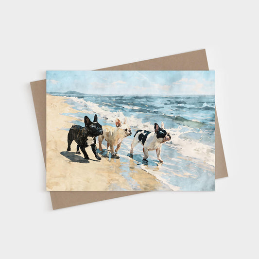 Three French Bulldogs walking together along the ocean shoreline under a cloudy blue sky, printed on a blank folded greeting card.