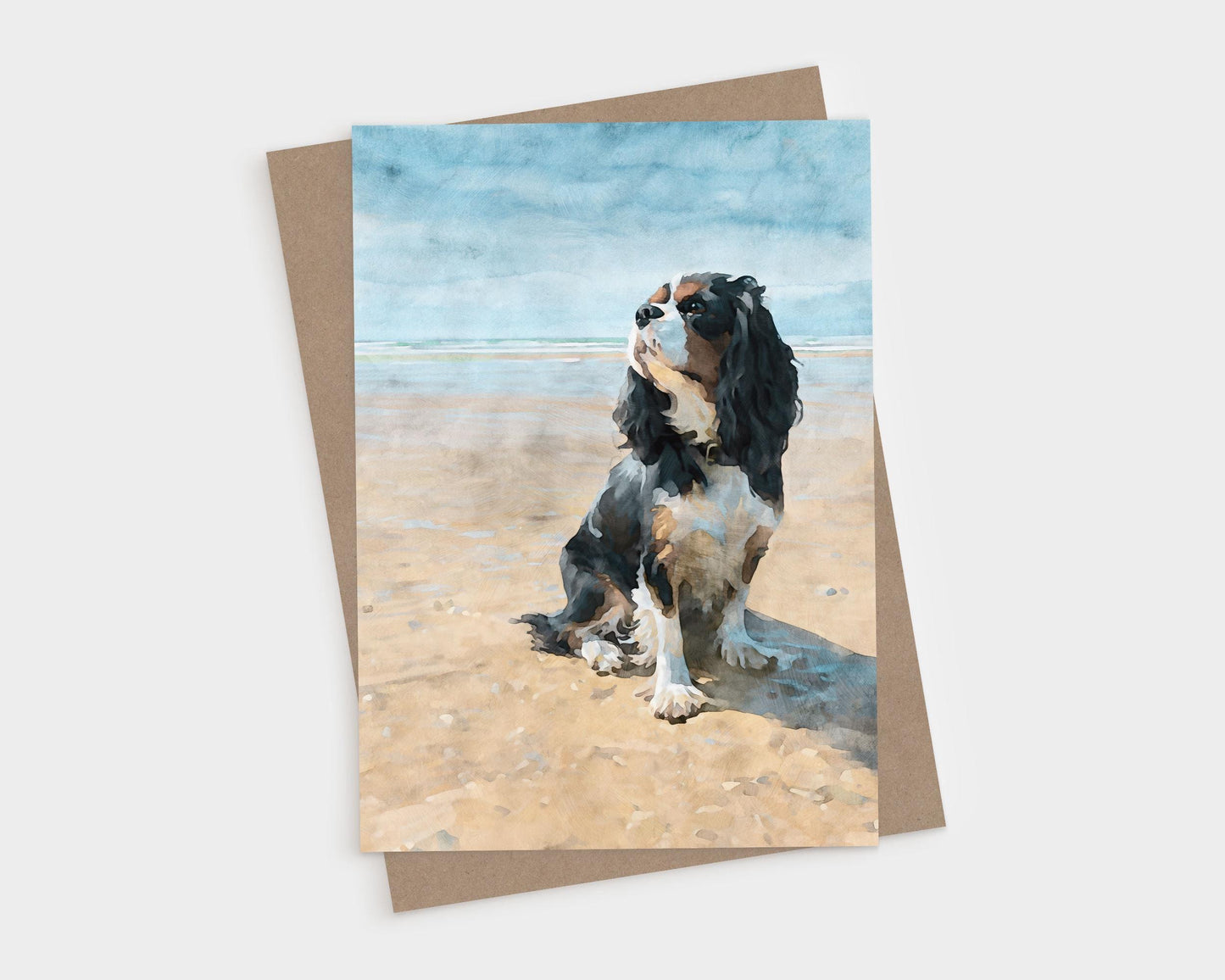 Tricolor Cavalier King Charles Spaniel sitting on a sunny beach, shown on a vertical blank folded greeting card with kraft envelope.
