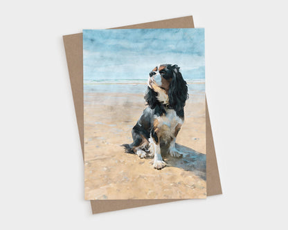 Tricolor Cavalier King Charles Spaniel sitting on a sunny beach, shown on a vertical blank folded greeting card with kraft envelope.