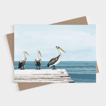 Three pelicans standing on a dock beside calm blue water, shown on a blank folded note card with kraft envelope.