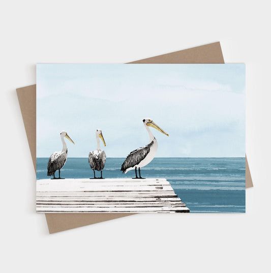 Three pelicans standing on a dock beside calm blue water, shown on a blank folded note card with kraft envelope.