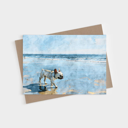 Cream French Bulldog standing in shallow beach water under a soft blue sky, shown on a blank folded greeting card with kraft envelope.